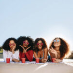Group of beautiful girls hanging out at the beach. Women making funny faces while lying on beach with cold drinks.