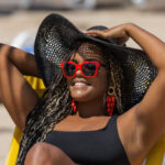 Close-up of an African-American woman on the beach lying on a deck chair wearing a black hat and sunglasses. Cheerful adult woman enjoying a summer day at the beach.