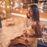 Beautiful young African woman in evening gown holding flute with champagne and smiling while spending time on night party